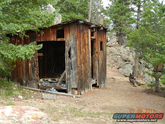 Dilapidated wooden shack with open doorway and broken walls, surrounded by trees and rocky terrain