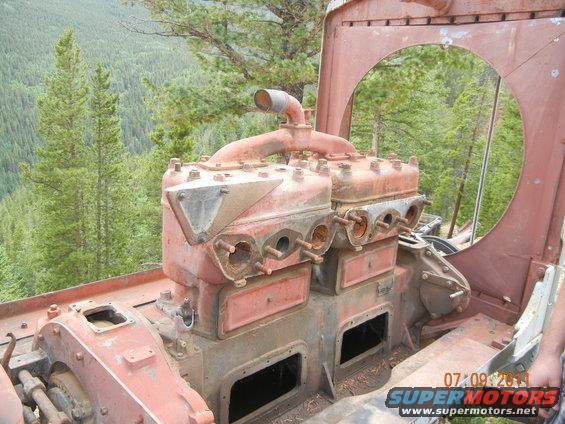 Close-up of a red engine block with exhaust pipes, set against a backdrop of trees. Watermark reads 'SUPERMOTORS.NET'