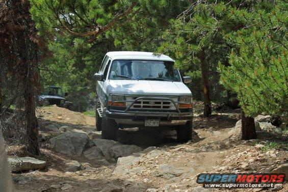 White SUV with brush guard driving over rocks on a dirt trail, another SUV visible in background