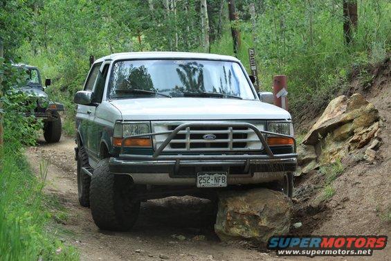 White Ford Bronco driving over a rock on a dirt trail, another Jeep is behind it