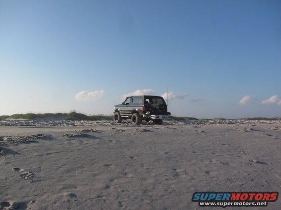 Dark 1995 Ford Bronco parked on a sandy beach with dunes and blue sky