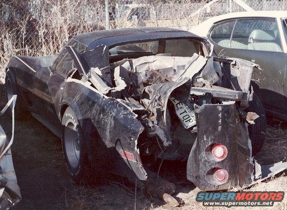 Rear end of a wrecked black 1976 Chevrolet Corvette with a Texas license plate reading 'LEE TOP'. Another car is visible behind it