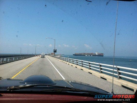 Dashboard of a red vehicle with a black hood scoop, seen from the interior looking out over a bridge with a cargo ship in the distance