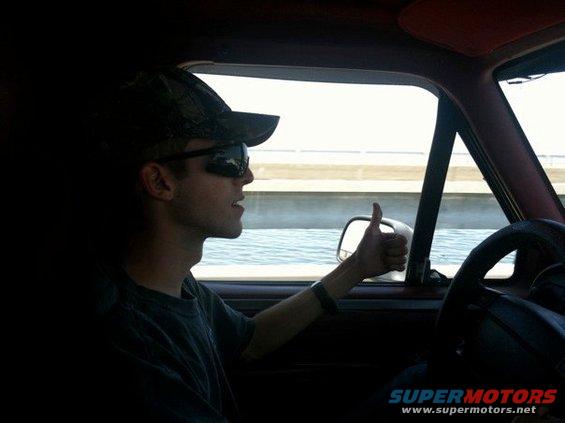Guy in camo hat and sunglasses gives a thumbs up while driving. Water and a bridge are visible outside