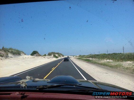 View from inside a vehicle looking down a road with sand dunes and vegetation on the side; two other vehicles are visible ahead