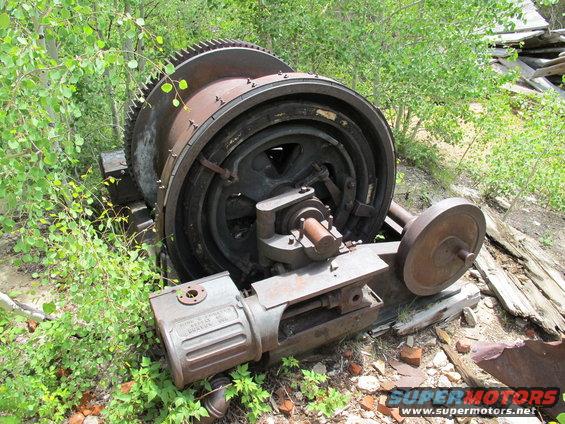 Large rusty industrial winch mechanism with a large gear and a smaller wheel, overgrown with vegetation