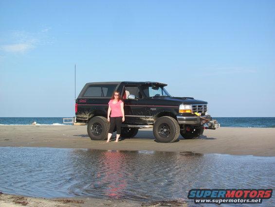 1995 Ford Bronco on a sandy beach with the ocean in the background. Woman in pink shirt stands beside it