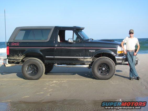 Black Ford Bronco on a sandy beach with a man standing by the front fender. The vehicle has mud splatters and a removable top