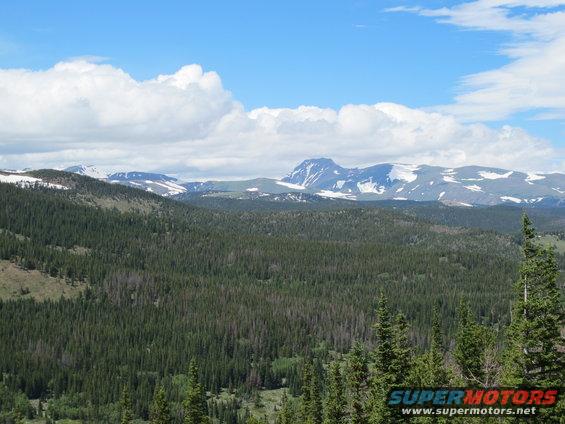 Mountain landscape with pine forest and snow-capped peaks under a blue sky with clouds. Watermark reads SUPERMOTORS