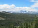 1994 Ford Bronco - A scenic view of a vast pine forest leading to snow-capped mountains under a bright blue sky with scattered clouds. The image includes a wa…