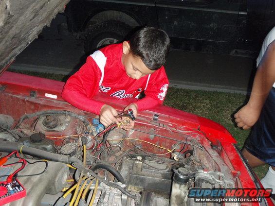 Red car engine bay with a boy working on wires, another person stands nearby