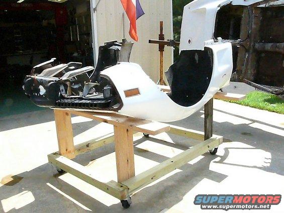 White car body section with exposed engine components, resting on a wooden stand