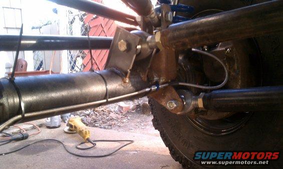 Close-up of a vehicle's rear axle with custom suspension components and brake lines. An angle grinder is on the ground nearby