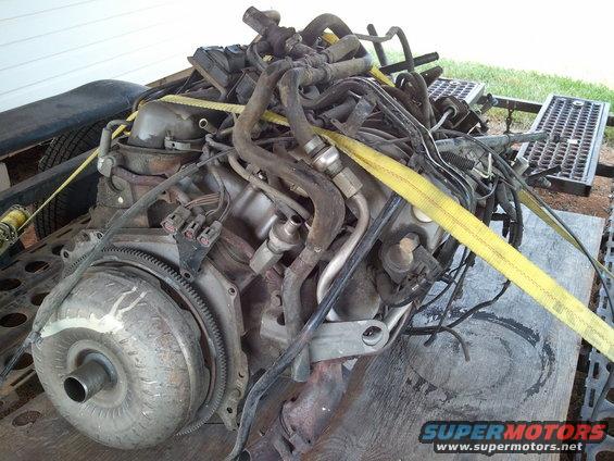 Engine and transmission assembly secured with yellow straps on a wooden surface, with a trailer in the background