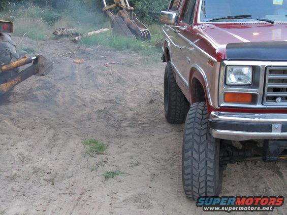 Red Ford Bronco with lifted suspension and large tires parked on dirt. Excavator in background