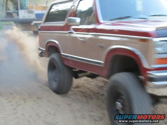 Red and tan Ford Bronco kicking up dust, with a construction vehicle in the background