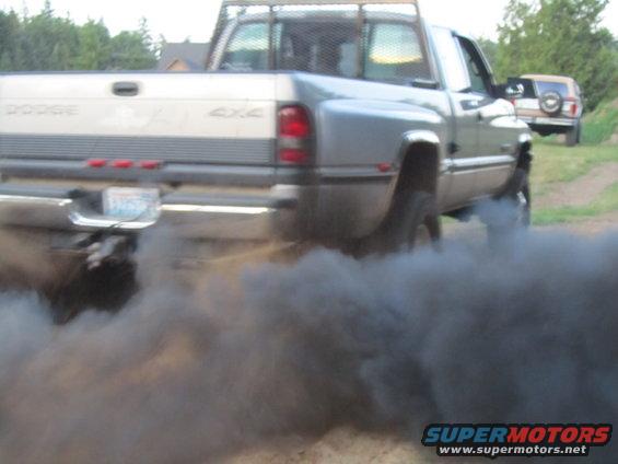 Dodge pickup truck rear view, emitting black smoke, with a second SUV in the background