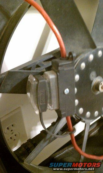 Close-up of an electric fan motor with a red wire and a clear fuse holder. A wall outlet is visible in the background