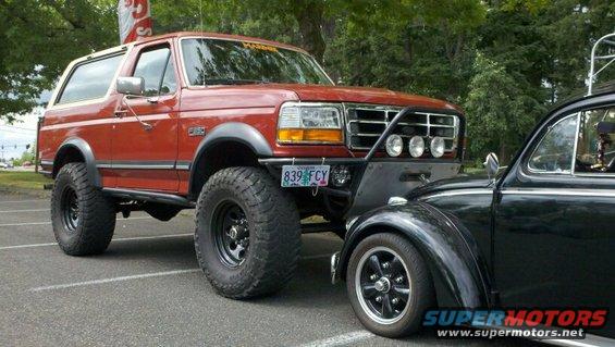 Red lifted Ford Bronco parked next to a black VW Beetle. Bronco has a custom bumper and lights