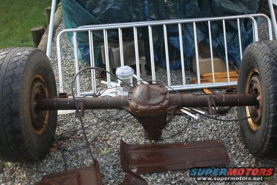 Rusty rear axle with two wheels, brake lines, and some metal trim pieces on gravel