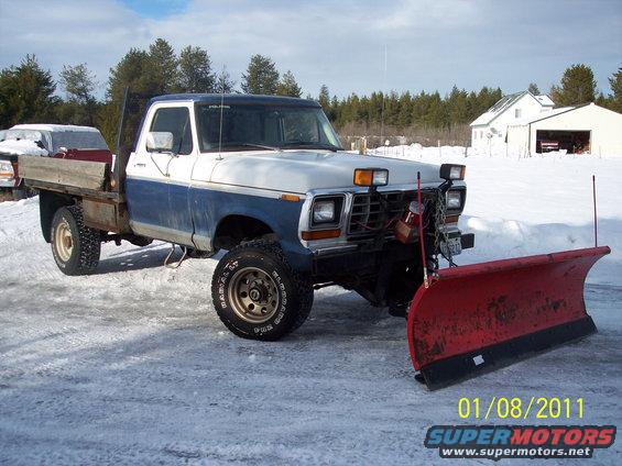 Flatbed Ford truck with a snow plow attached, parked in the snow. Another vehicle is partially visible behind it