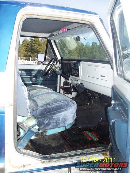 Pickup truck interior with bench seat, dashboard, and steering wheel. Floor mat has text