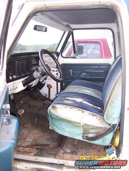 Interior view of a worn truck cab with striped seats and a floor mat. Another truck is visible outside