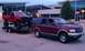 1994 Ford F-250 - A maroon SUV tows a red pickup truck on a trailer, with a large building in the background.
