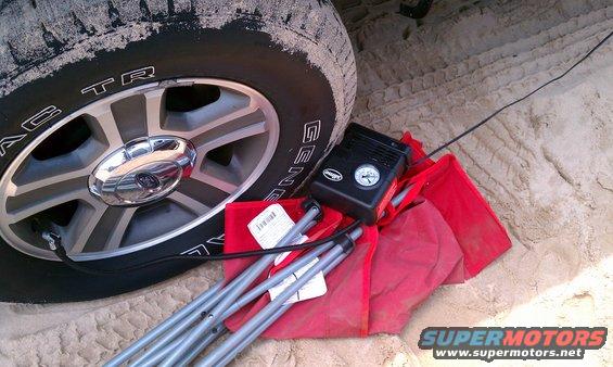 Close-up of a tire with 'GENERAL TIRE' visible, next to a black air compressor and red bag with metal poles on sandy ground
