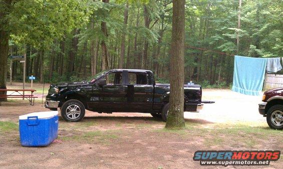 Black crew cab pickup truck parked at campsite with blue cooler nearby. Another truck is partially visible on the right