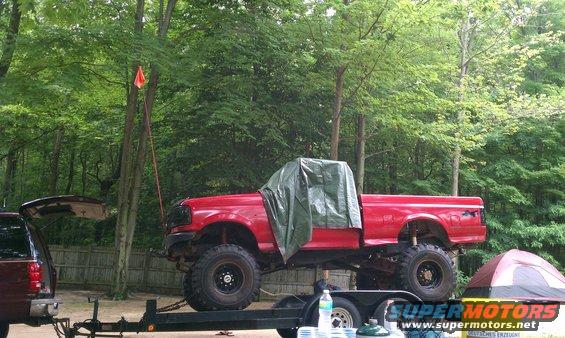 Red pickup truck with a tarp over the cab, lifted on a trailer. A dark SUV is partially visible on the left