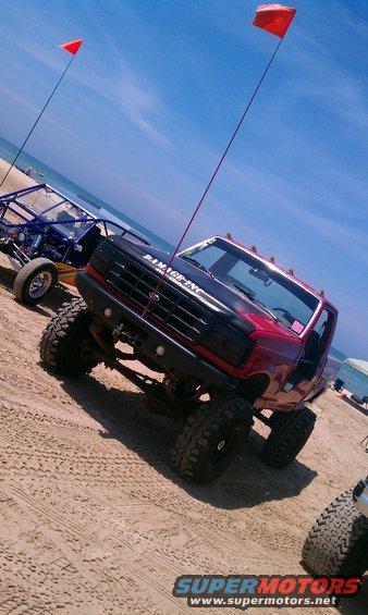 Red 1994 Ford F-250 with large tires on sand, "DAMAGE INC" on hood