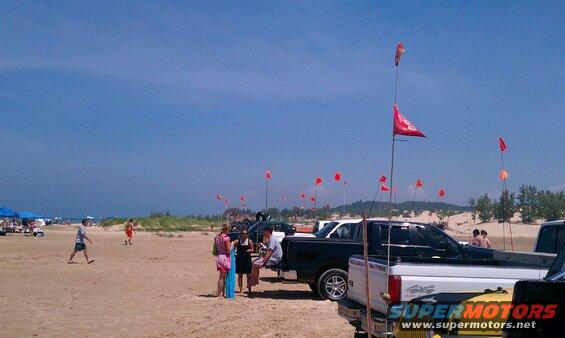 Trucks parked on a sandy beach with people milling around and flags on poles