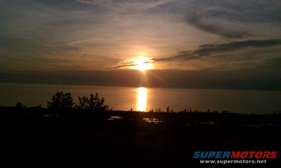 Sunset over a body of water with silhouetted trees and land in the foreground. Watermark reads SUPERMOTORS