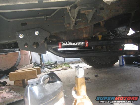 Underbody view of a vehicle showing a black bracket and a steering stabilizer with 'LAMBURE' text