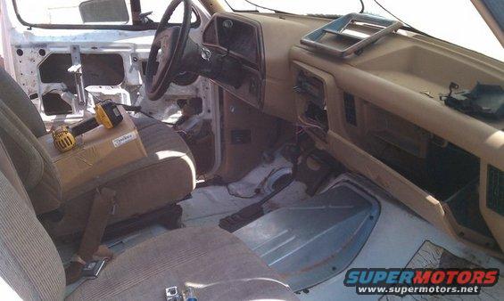 Interior of a 1990 Ford Bronco in disrepair, showing disassembled door panels, exposed wiring, and a drill on the passenger seat