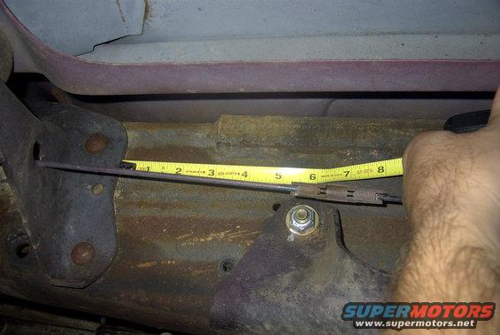 Close-up of a hand holding a tape measure against a rusty metal cable and bracket on a vehicle underside