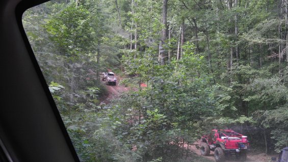 Two off-road vehicles driving on a wooded dirt trail. One is red and in the foreground, the other is lighter colored and further up the trail