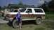 1989 Ford Bronco - Two trucks are parked outside cabins, one is a white and brown Ford Bronco covered in mud.