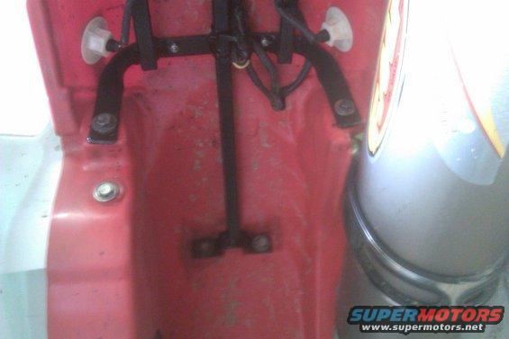 Close-up of a red plastic rear fender mount with black metal brackets and bolts. Part of a silver tank is visible on the right