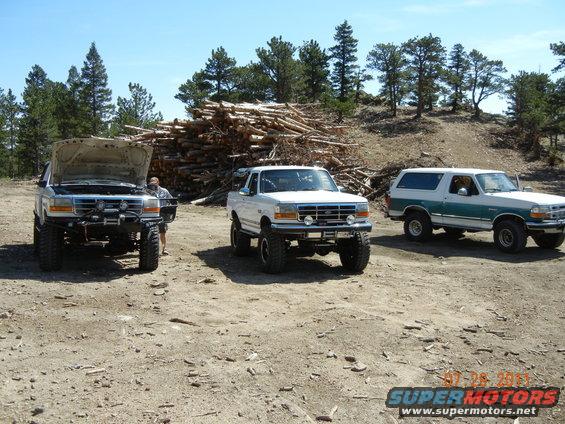 Three lifted Ford Broncos parked in a dirt lot. One has its hood open. A pile of logs is in the background