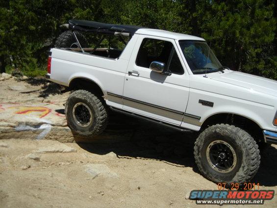 White Ford Bronco with large tires climbing over rocks. Has a soft top over the rear