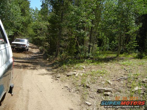 White 1996 Ford Bronco driving through mud on a dirt road, seen from another vehicle