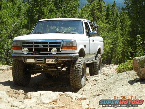 White 1996 Ford Bronco with large tires on a rocky, wooded trail