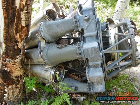 large metal engine component with pipes and a cage-like structure, leaning against a tree