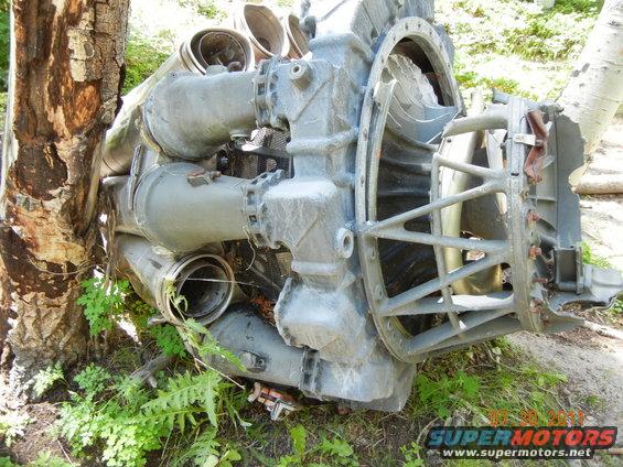 Gray engine component with multiple pipes and a large fan housing, resting on the ground near a tree