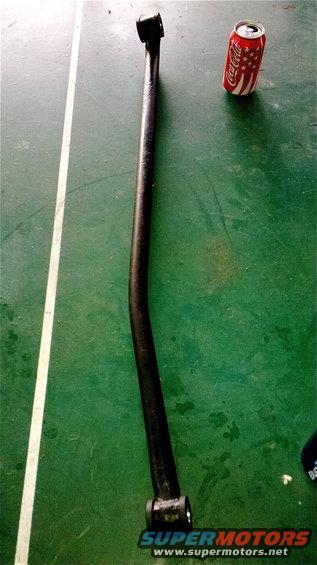 Black suspension control arm with rubber bushings on a green surface next to a Coca-Cola can