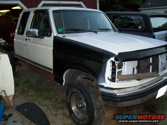 White Ford Bronco with black fender, undergoing repairs. Another dark SUV is parked behind it