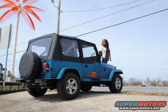 Blue 1991 Jeep YJ with black soft top and spare tire on rear, parked on pavement near water