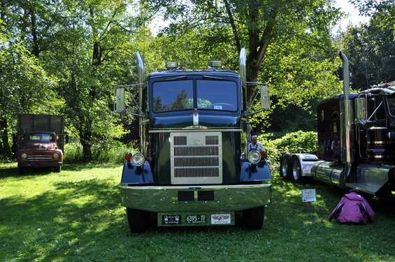 Front view of a dark green semi-truck with chrome grille and stacks. A red truck is parked behind it to the left